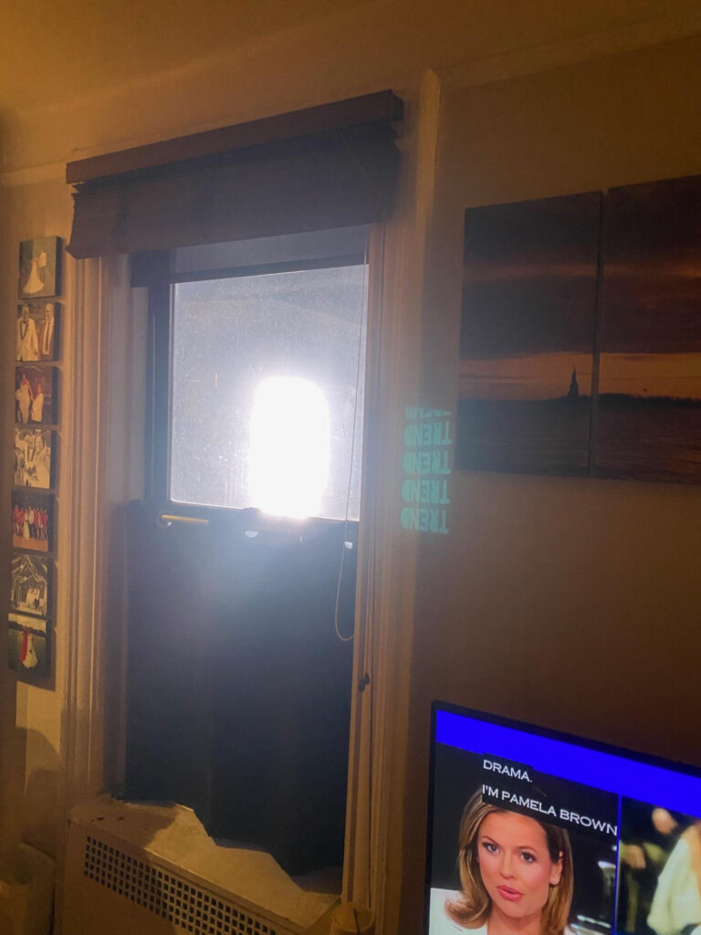 Interior of a living room at 205 West 54th Street at night, the window filled with light from the sign and the ad’s text projected on the wall